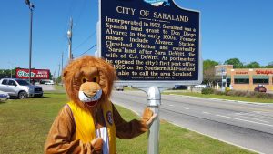Leo, the Saraland Lions Club mascot, standing at the historical marker.